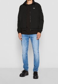 Black zip-up bomber jacket with a high collar, paired with a black T-shirt, light blue jeans, and black lace-up shoes.