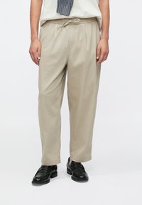 Pantalon beige en mélange de coton avec une ceinture élastique et un cordon de serrage, doté d'une coupe décontractée et de poches latérales, porté avec des chaussures noires.