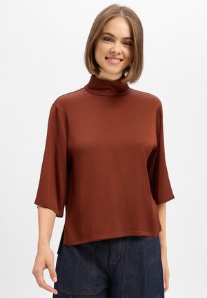 Jeune femme souriante portant un haut marron à col montant avec des manches trois-quarts et un jean bleu foncé, sur un fond clair uni.