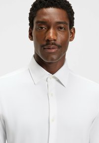 Chemise blanche à col, en tissu texturé avec fermeture à boutons, présentant une coupe ajustée et un léger brillant.