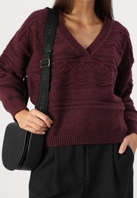 Vinröd stickad tröja med V-ringning och strukturerat mönster. Svart axelremsväska med logotyp, bärs över svarta byxor.