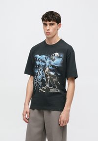 Juoda grafikos marškinėliai su vilko, žaibo ir mėnulio dizainu mėlynos ir baltos spalvos. Trumpomis rankovėmis, laisvo silueto, audinys atrodo lygus.