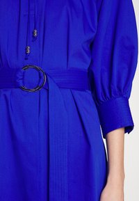 Robe bleu vif avec manches ballon 3/4, large poignet, ceinture nouée avec boucle circulaire argentée et accents de perles argentées sur les liens avant.