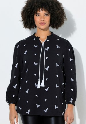 Kobieta o kręconych włosach, ubrana w czarną bluzkę z długim rękawem z białymi haftowanymi sercami i białą frędzlą przy dekolcie.