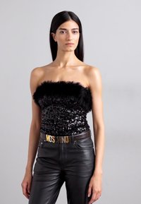 Čierny top bez ramienok so sequinmi a pierkovým lemovaním, skombinovaný s čiernymi koženými nohavicami a zlatým opaskom s logom Moschino.