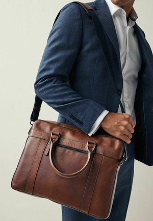 Man in navy suit and white shirt carrying brown leather briefcase with shoulder strap against plain background.