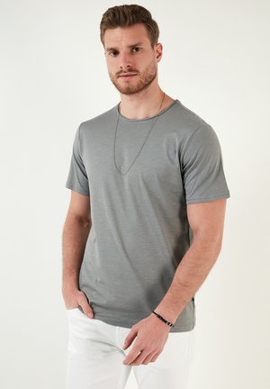 Grå kortærmet t-shirt med rund hals, glat tekstur og subtil glans. Båret med hvide bukser; en lang halskæde synlig.