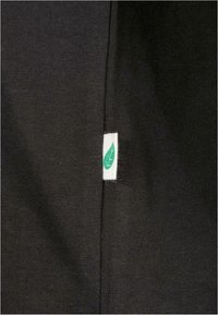 Fekete szövet finom bordázott mintázattal, kis fehér címkével, zöld levél logóval a varrásnál. Egyszerű, letisztult design.