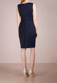 Marineblauw mouwloos jurkje met een getailleerde snit, voorzien van een naaddetail ter hoogte van de taille en een ritssluiting aan de achterkant. Beige hoge hakken.