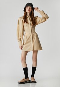Beige Kleid mit langen Puffärmeln und Kragen, kombiniert mit schwarzen kniehohen Socken und braunen Schuhen. Das Model trägt einen schwarzen Bucket Hat.