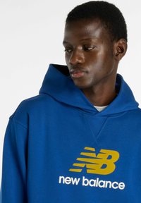 Blå luvtröja med framfickan i kengurustil, med en stor gul och vit "new balance"-logotyp. Mjuk tygstruktur.