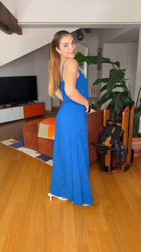 Νεαρή γυναίκα με μακρύ μπλε φόρεμα και λευκά τακούνια ποζάρει χαμογελαστή σε ένα σαλόνι με ξύλινο πάτωμα, φυτά και έπιπλα.