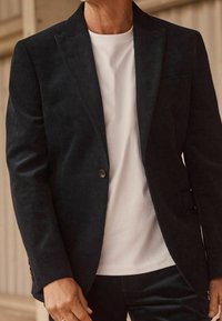 Homme portant un blazer en velours côtelé noir et un t-shirt blanc à col rond, debout à l'intérieur avec la main détendue le long du corps.