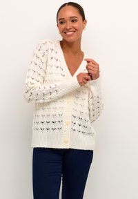 White knitted cardigan with a V-neck, button closure, and openwork pattern, featuring ribbed cuffs and hem. Worn over dark jeans.