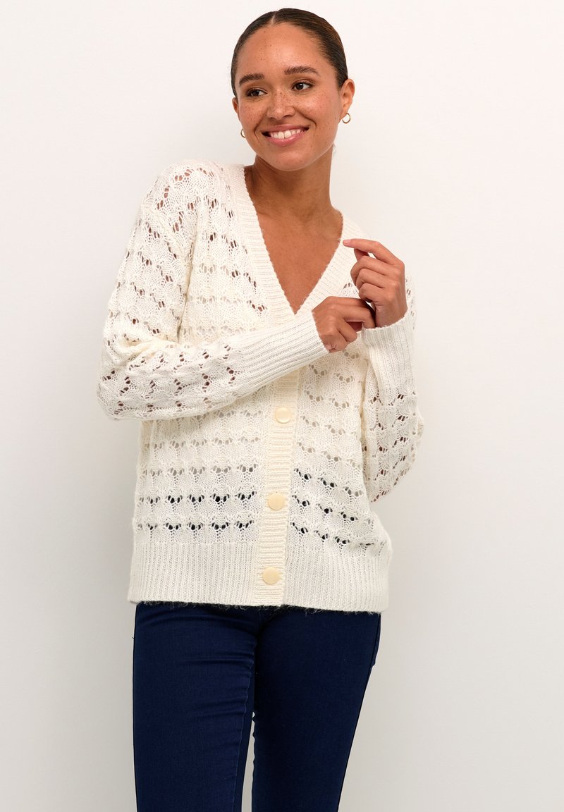 White knitted cardigan with a V-neck, button closure, and openwork pattern, featuring ribbed cuffs and hem. Worn over dark jeans.