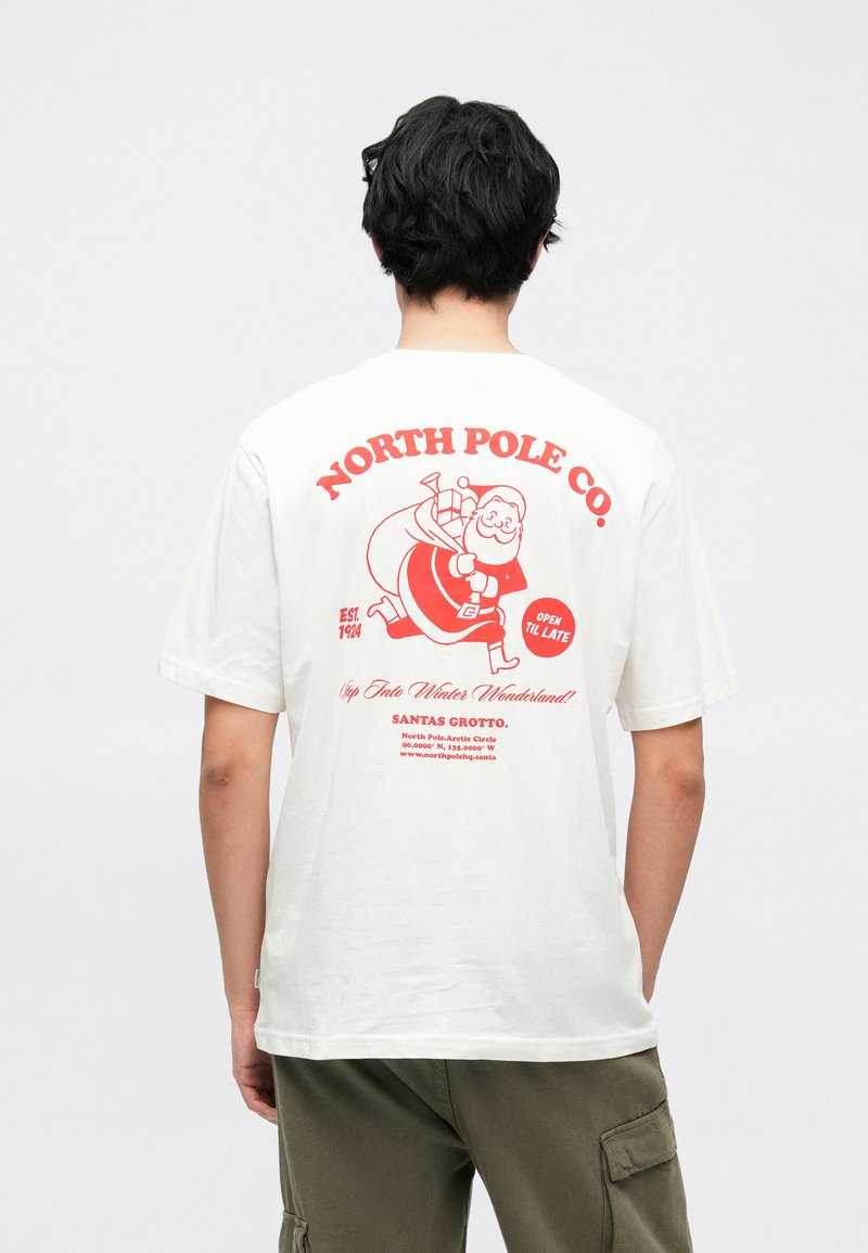 Hvid bomuld t-shirt med rød grafik af julemanden med en pose og teksten "NORTH POLE CO." på ryggen. Korte ærmer, løs pasform.