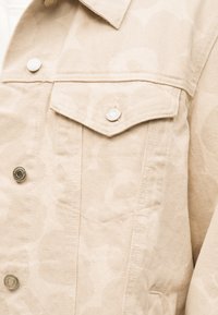 Μπεζ τζιν μπουφάν με τσέπη στο στήθος με καπάκι και ασημένια κουμπιά, που δείχνει λεπτομερή υφή ύφασματος και ραφές.