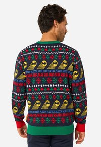 Kampsun tumeda taustaga, millel on värvilised jõulupärlid, sealhulgas kollased batmanimärgid, punased ja rohelised aktsendid ning soonikkäised.