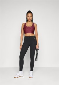 Kvinne i rød mønstret sports-bh og svarte leggings som holder et svart hoppetau, stående i hvite sneakers mot en enkel bakgrunn.
