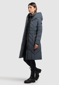 Dolga podložena jakna temno sive barve s kapuco, s pikčasto vzorčasto z diamantnim šivom, sprednja zadrga in stranski žepi; nošena s črnimi škornji.