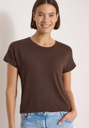 Frau mit kurzen braunen Haaren, die lächelt, trägt ein braunes, strukturiertes Kurzarmshirt und hellblaue Jeans mit einer Hand in der Tasche.