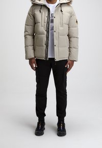 Doudoune beige avec une capuche doublée en fausse fourrure, fermeture éclair, et deux poches avant, associée à un pantalon cargo noir et des chaussures noires.