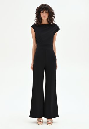 Femme aux cheveux bouclés et foncés, portant une combinaison noire sans manches à jambes larges et des sandales ouvertes dorées, debout devant un fond blanc.