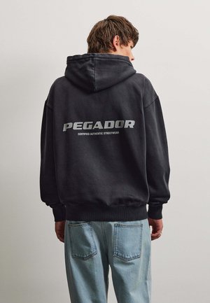 Giovane uomo con capelli castani indossa una felpa nera con il testo "PEGADOR Certified Authentic Streetwear" sulla schiena e jeans azzurri, rivolto verso il muro.