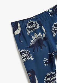 Navyblauwe katoenen leggings met dinosaurustekenen in crème en donkerblauw, en speelse teksten "ROAR!" en "RUDE!" in spraakballonnen.