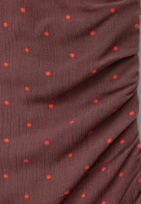 Tissu marron avec une apparence texturée, comportant des pois rouges éparpillés sur une surface légèrement froncée.