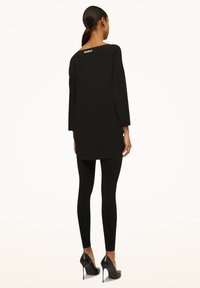 Sort tunika med trekvartærmer, med en lige pasform og diskret logodetalje bagpå. Kombineret med sorte leggings og hæle.