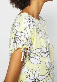 Blomsterprint top i lysegul med sorte kanter. Har korte ærmer med samlede detaljer og bånddetaljer ved ærmerne.