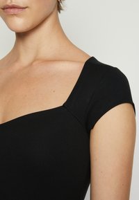 Svart topp med korta ärmar, fyrkantig halsringning, figursydd design, slät textur och subtila sömnadsdetaljer runt halsringningen och ärmarna.