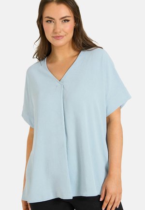 Vrouw met een lichtblauw, los vallend V-hals topje met korte mouwen, staand tegen een effen witte achtergrond, naar voren gericht met een subtiele glimlach.