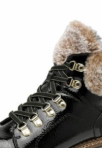 Fekete lakkbőr csizma műszőrme gallérral, arany fémlyukakkal és fekete fűzőkkel, melyeken fehér díszítések találhatók. A texturált felület mélységet ad.