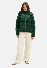 Groene pufferjack met een hoge kraag, ritssluiting en gewatteerde textuur, gecombineerd met lichtbeige wijduitlopende broek en casual schoenen.