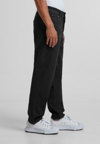 Jean en denim noir avec une coupe droite, dotés de poches latérales et d'une braguette zippée. Portés avec des baskets blanches. Texture lisse et design minimal.