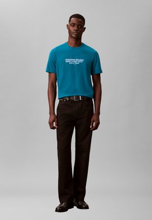 Mann trägt ein türkisfarbenes Calvin Klein T-Shirt, schwarzen Gürtel, schwarze Hose und schwarze Schuhe und steht vor einem schlichten grauen Hintergrund.