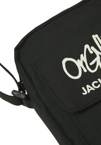 Bolso de tela negra con cierre de cremallera, texto de marca blanco prominente y superficie texturizada. Forma rectangular compacta con correa ajustable.