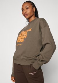 Hnedý oversized sveter s oranžovým textom „ZDRAVIE & TELLO & MYSEĽ & DUŠA.“ Zmes bavlny, rebrované manžety a okrúhly výstrih.