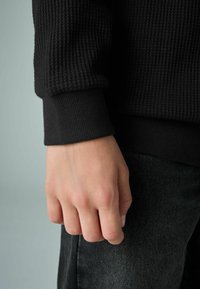 Čierny rebrovaný rukáv sveter s textúrovaným povrchom, kombinovaný s tmavými džínsami. Viditeľná ruka zaťatá v päsť, ukazujúca detaily tónu pleti.