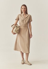 Beige, kurzärmliges, kragenloses Kleid mit einem Gürtel in der Taille und weicher Textur. Das Outfit wird mit beigen Sneakers und einer passenden Tragetasche kombiniert.