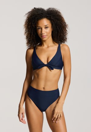 Keharad naine, kes kannab tumesinist bikiinid esiknodiga peal ja kõrge vöökohaga bottoms, seisab ühevärvse hele tausta ees.