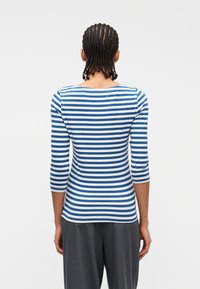 Άτομο με πλεγμένα μαλλιά που φοράει μπλε και λευκή ριγωτή μπλούζα με μακριά μανίκια και σκούρο γκρι παντελόνι, γυρισμένο προς τα πίσω σε απλό φόντο.