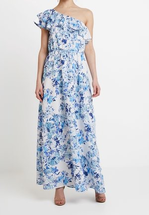 Frau trägt ein langes, einseitiges weißes Kleid mit blauem Blumenmuster und gerafftem Ausschnitt, kombiniert mit beigen offenen Schuhen.