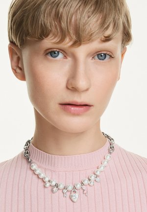 Collana con catena d'argento e perle bianche rotonde, con ciondoli a forma di cuore e chiave, indossata sopra un maglione rosa chiaro a coste.