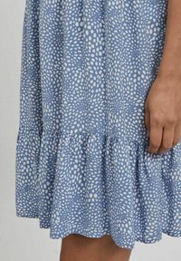 Blå klänning i lätt tyg med ett vitt abstrakt prickmönster, volangkant och avslappnad passform. Synlig hand som vilar på tyget.