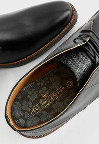 Zapatos de piel negra con cordones que presentan un upper suave, detalle texturizado en el talón y una plantilla con estampado floral y letras doradas.