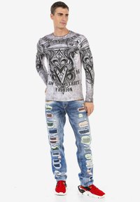 Langärmliges Grafikshirt in Grau mit schwarzen, eleganten Designs; kombiniert mit abgetragenen Jeans mit buntem Patchwork. Rote Sneakers runden den Look ab.