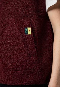 Bordo flis prsluk s teksturiranom površinom, s prednjim džepom i malom oznakom u zelenoj i žutoj boji s crvenim naglaskom u obliku srca.